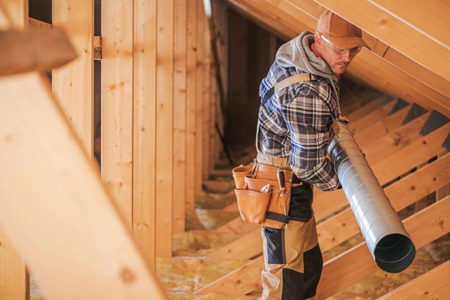 Ouvrier transportant un tuyau de ventilation dans un grenier en bois.