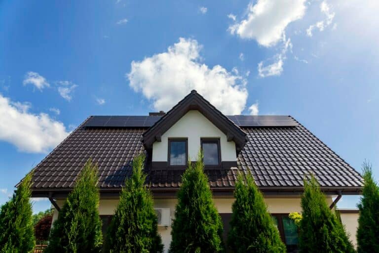 Vue d'une maison avec une toiture noire et des panneaux solaires installés sur le toit, entourée de verdure et sous un ciel bleu avec des nuages blancs.