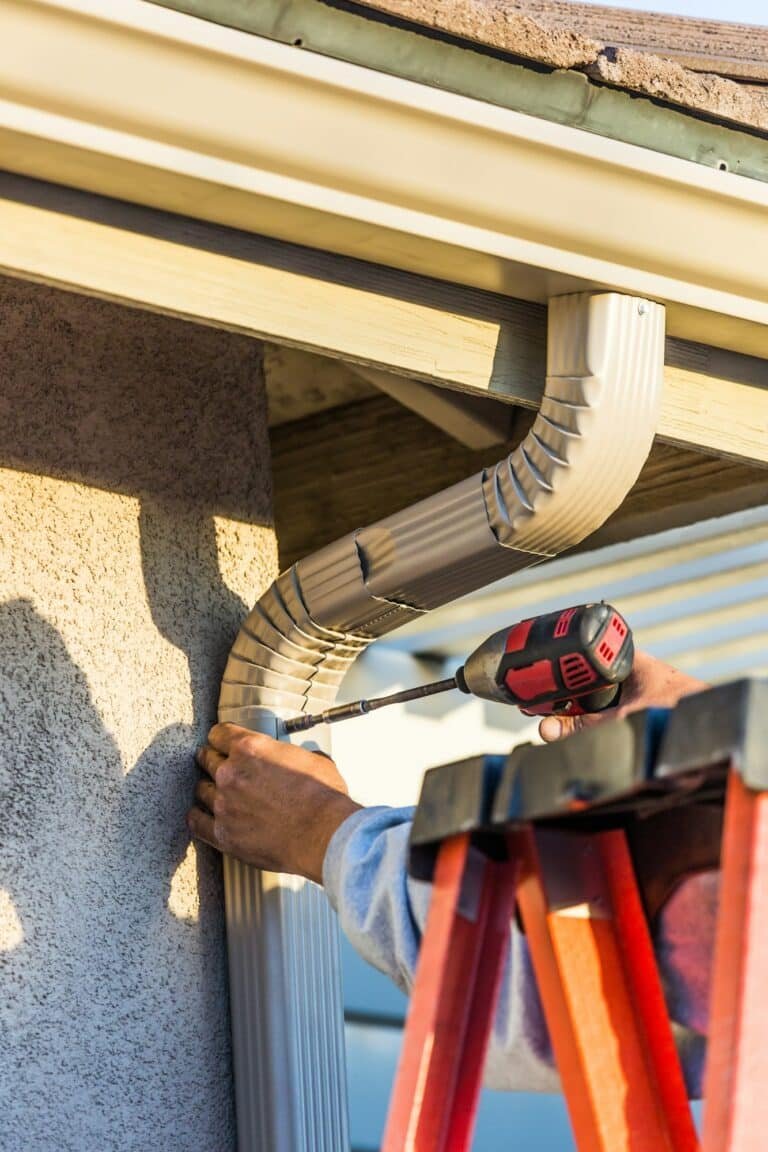 Gros plan d'un travailleur installant une gouttière sur le côté d'une maison à l'aide d'une perceuse, debout sur une échelle rouge.